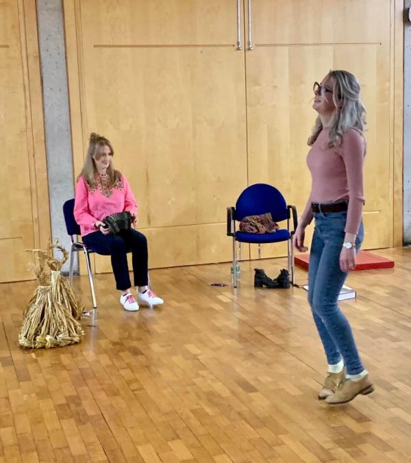 Women with blonde hair wearing a pink top and blue jeans dances to accordion music which is being played by woman with blonde hair wearing a pink top and black jeans sitting down 