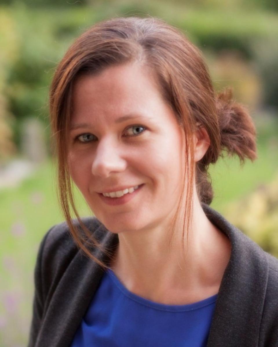 woman with brown hair in a black jacket and blue blouse against green grass and bushes