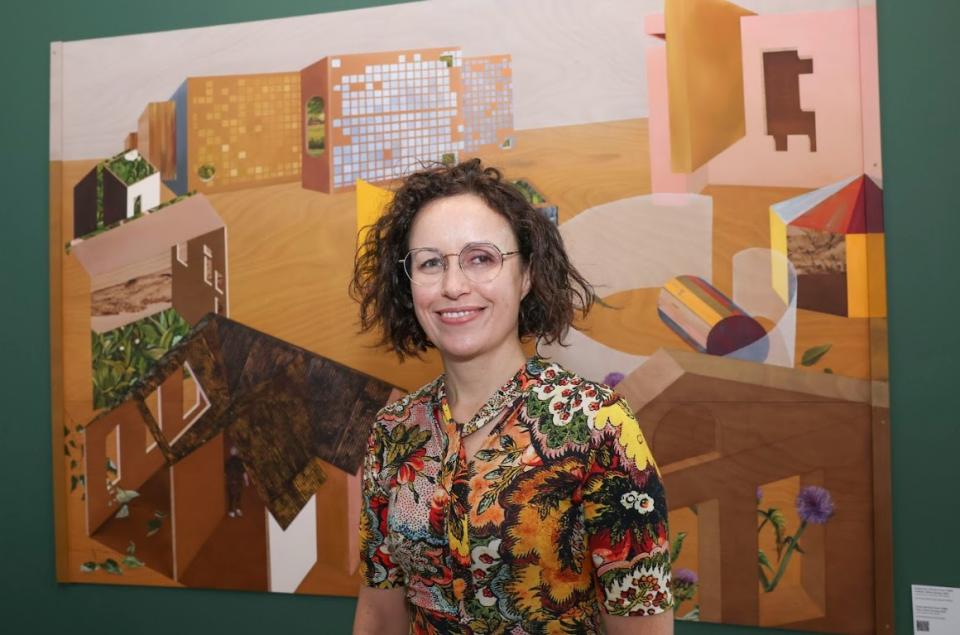 Artist Deirdre Frost, a women with brown curly hair and glasses stands in front of her artwork - Spreag. A painting which includes buildings from across the three campuses of DCU