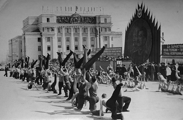 Shows a patriotic performance by dancers in Soviet Russia, decal showing Stalin and Lenin in the background