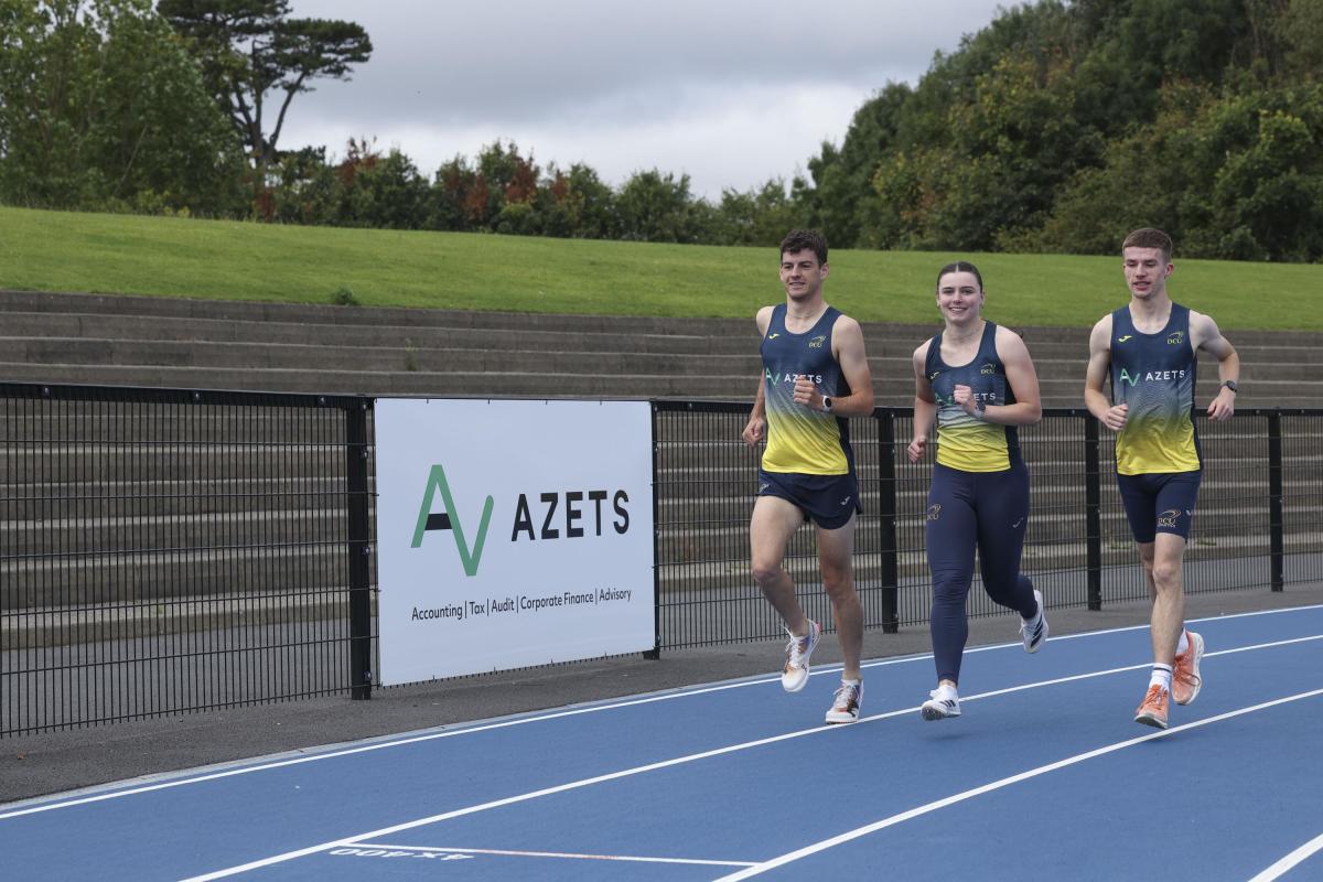 Three DCU athletes running past an Azets sign.