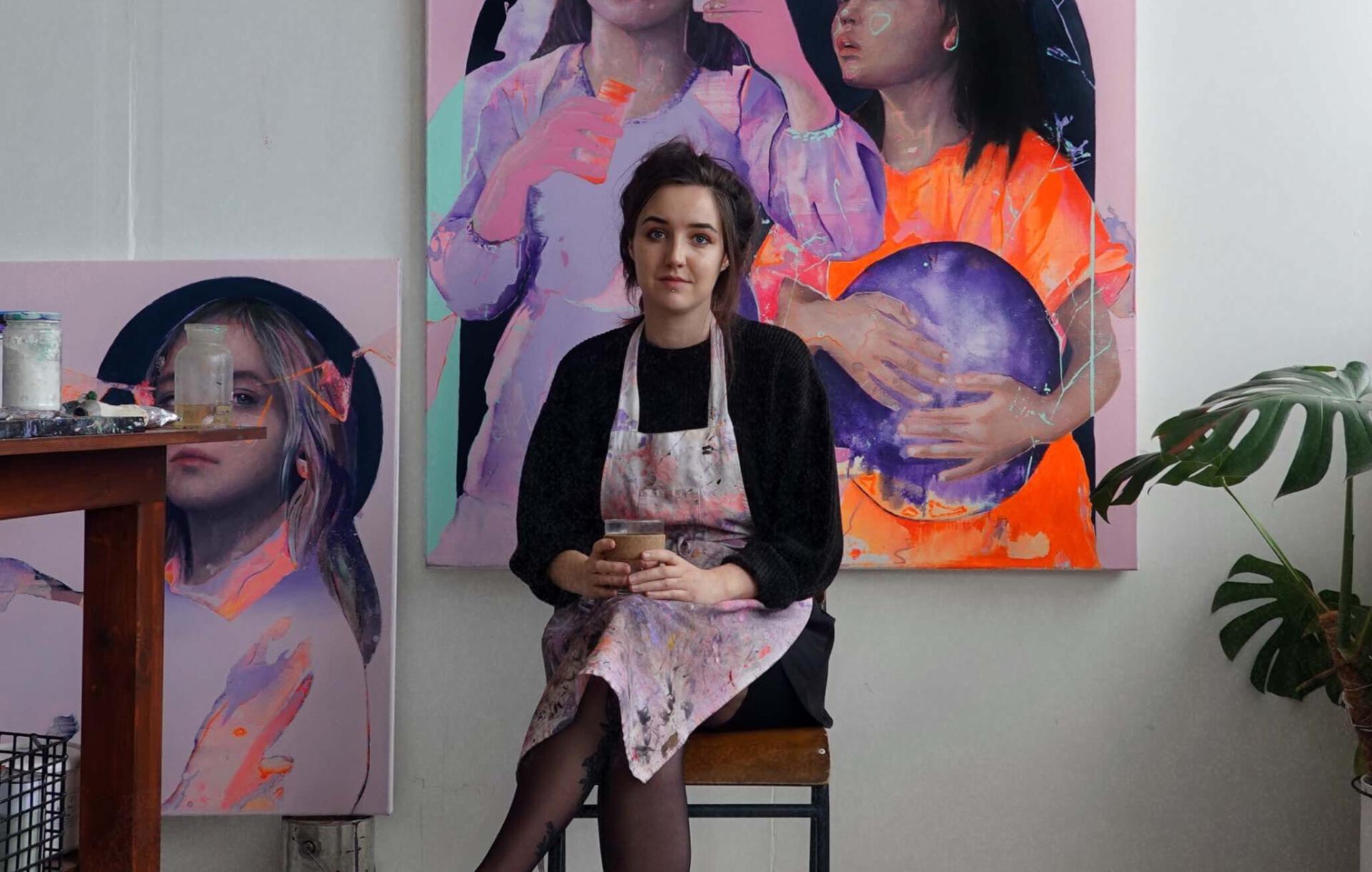 Artist Alana Barton, a girl with long brown hair tied up wearing a grey jumper sits in her studio