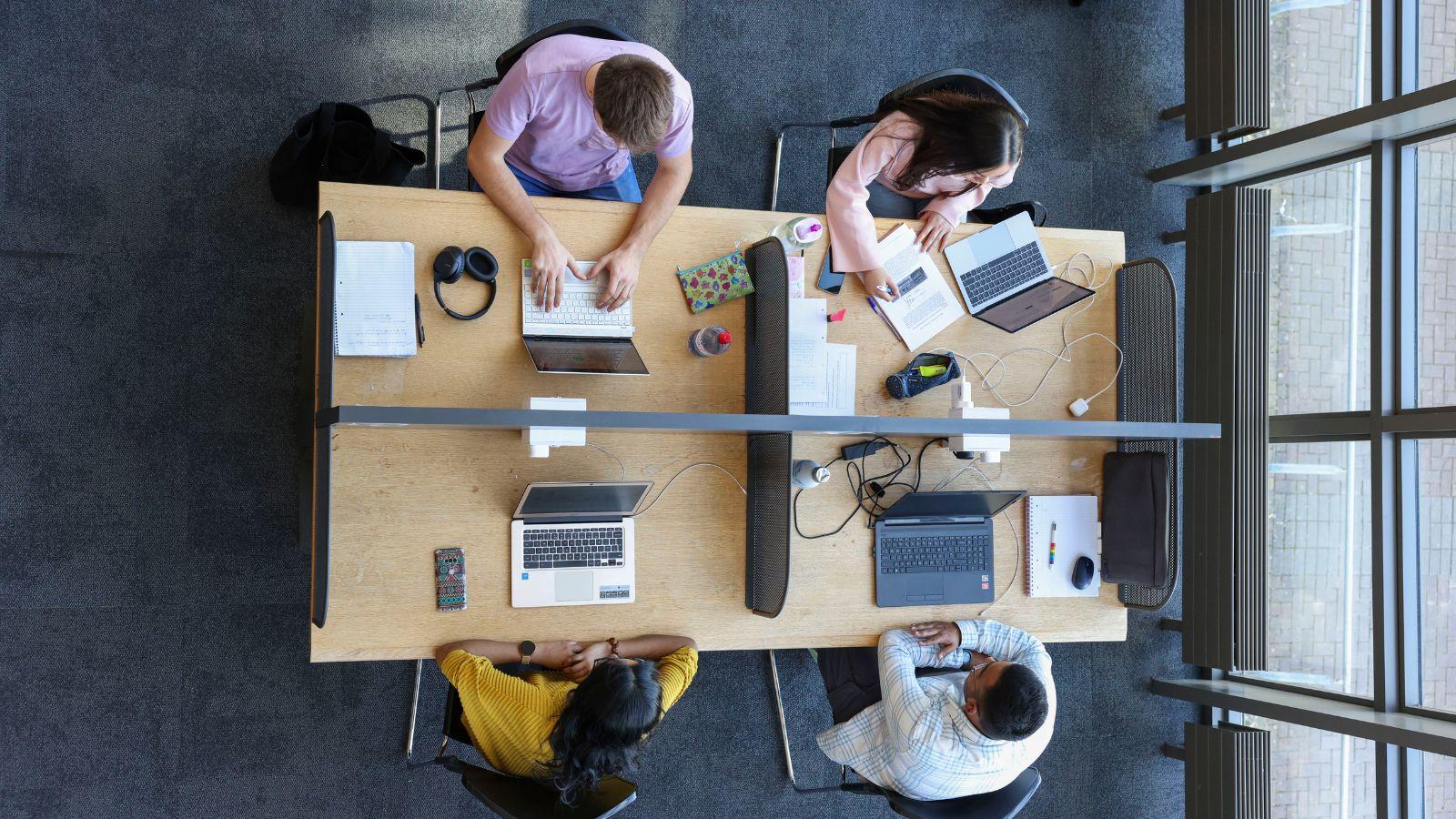 overhead shot of 4 students sitting at a quad of desks. Around them are various accessories to study: latpops, books, notepads, pens