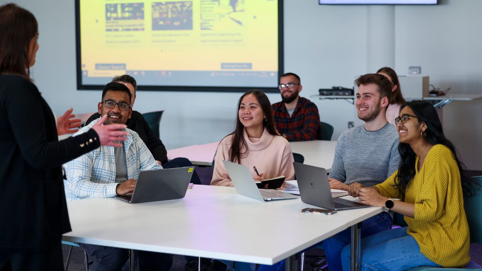 Students seated at a table looking up at a woman standing to the left of the picture