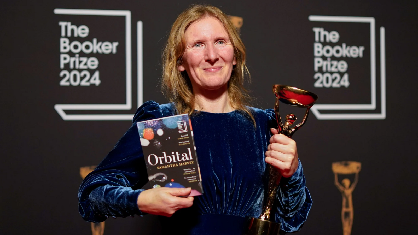 A woman stands holding a book and an award. A logo with The Booker Prize 2024 is in the background. 