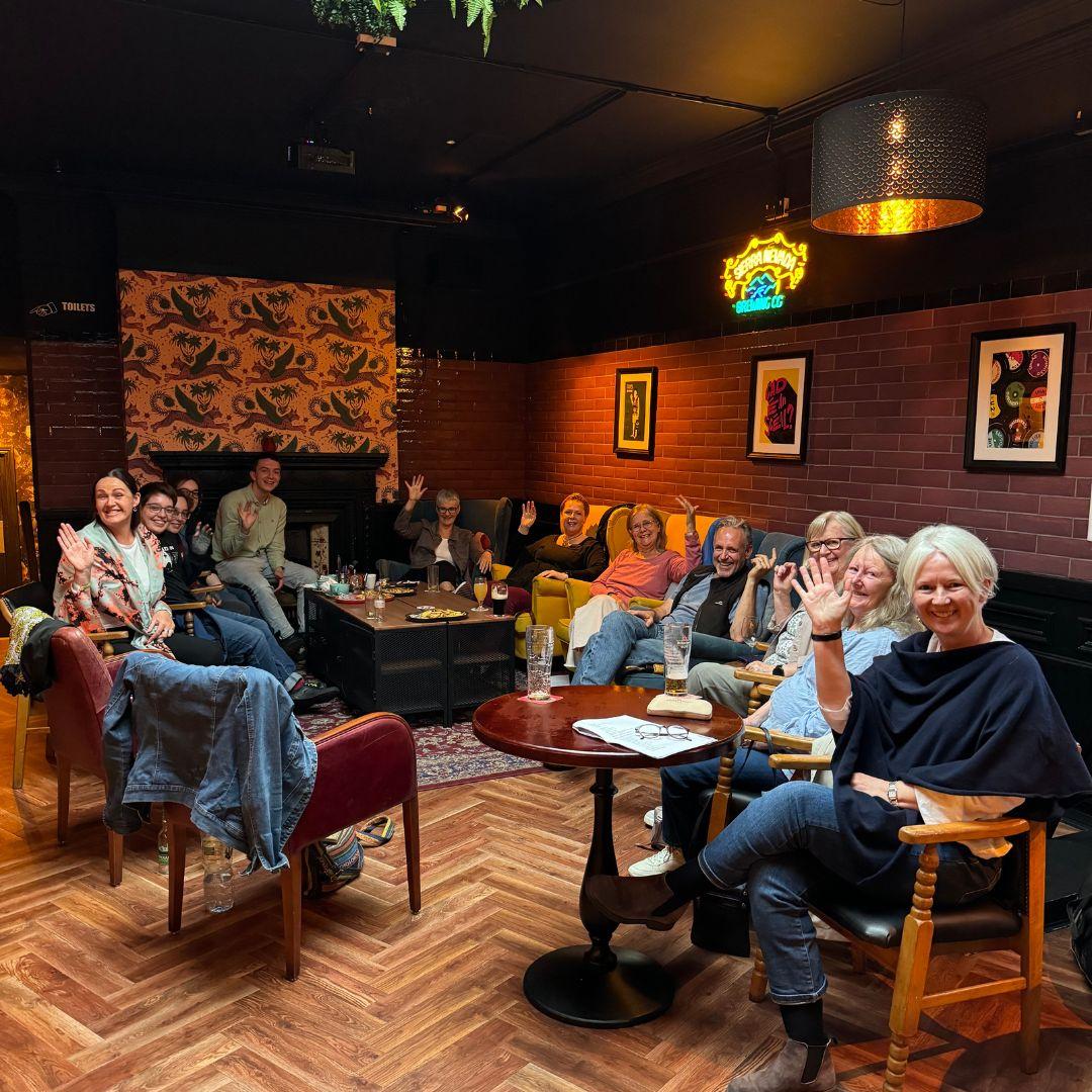 Group of people sitting in a pub facing the camera smiling and waving 